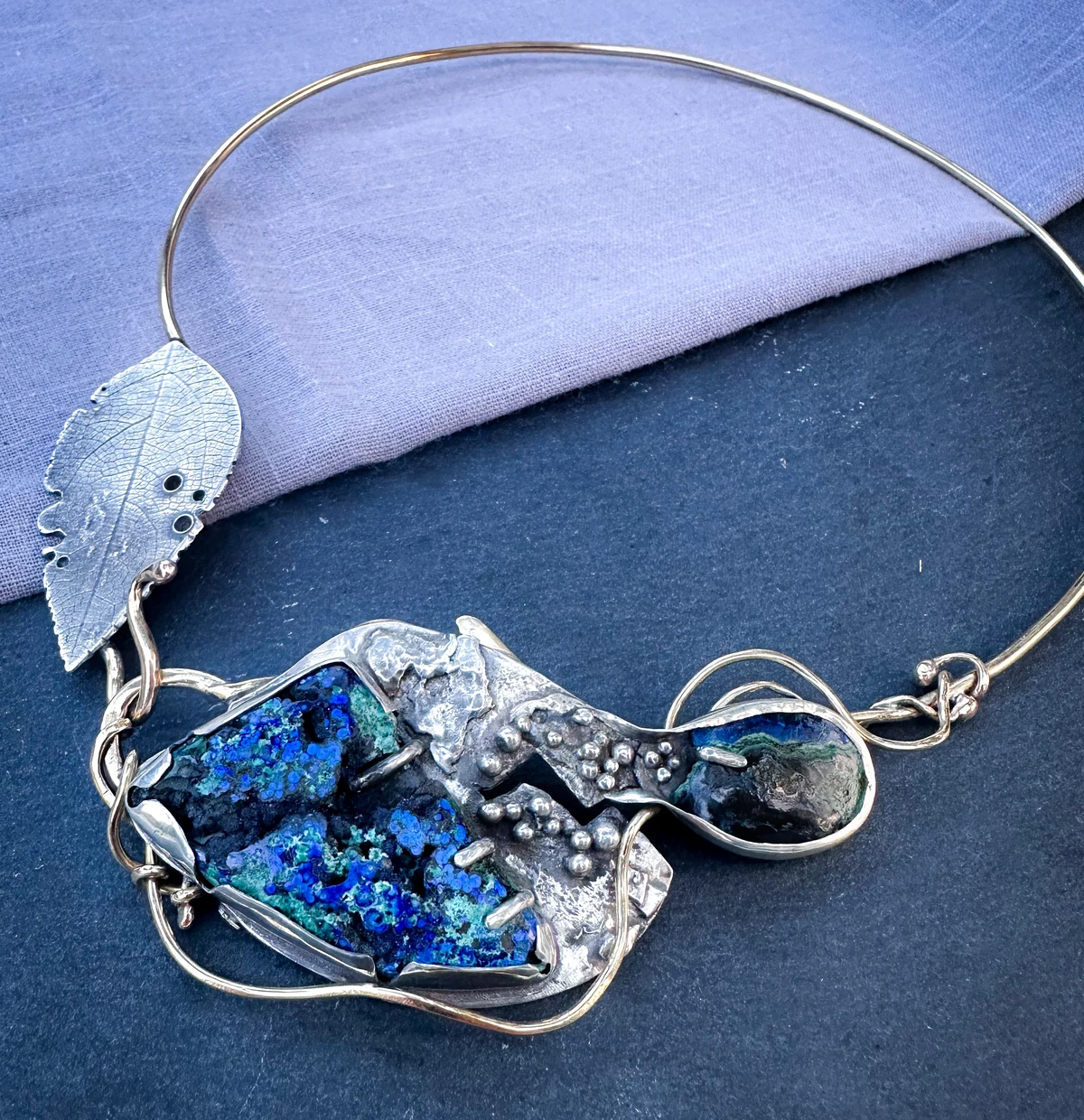 Beyond: an asymmetric necklace with a leaf-shaped metal element, a blue-green druse, a small oval stone and a delicate neckwire.